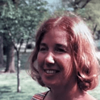 Color snapshot of a smiling woman with red hair outdoors among trees.