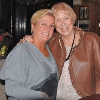Two smiling women, one blond in gray and one with red hair in a leather jacket, at a restaurant.